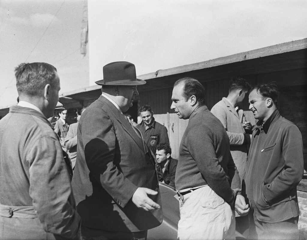 Fangio (centre, in the jumper) with the legendary Mercedes team manager Alfred Neubauer (in the hat) at the 1955 Dutch Grand Prix.
